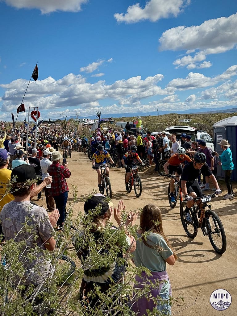 Riders launching from the start of 24 Hours in the Old Pueblo 2026 on the dirt road course at Willow Springs Ranch, surrounded by cheering spectators