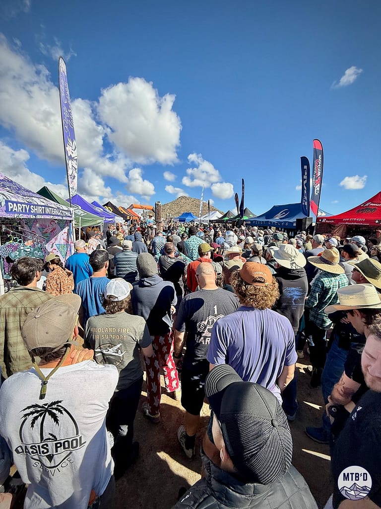 Riders gathered for the pre-race captains meeting at 24 Hours in the Old Pueblo, surrounded by vendor tents in the expo area near Tucson with desert hills and clouds in the background
