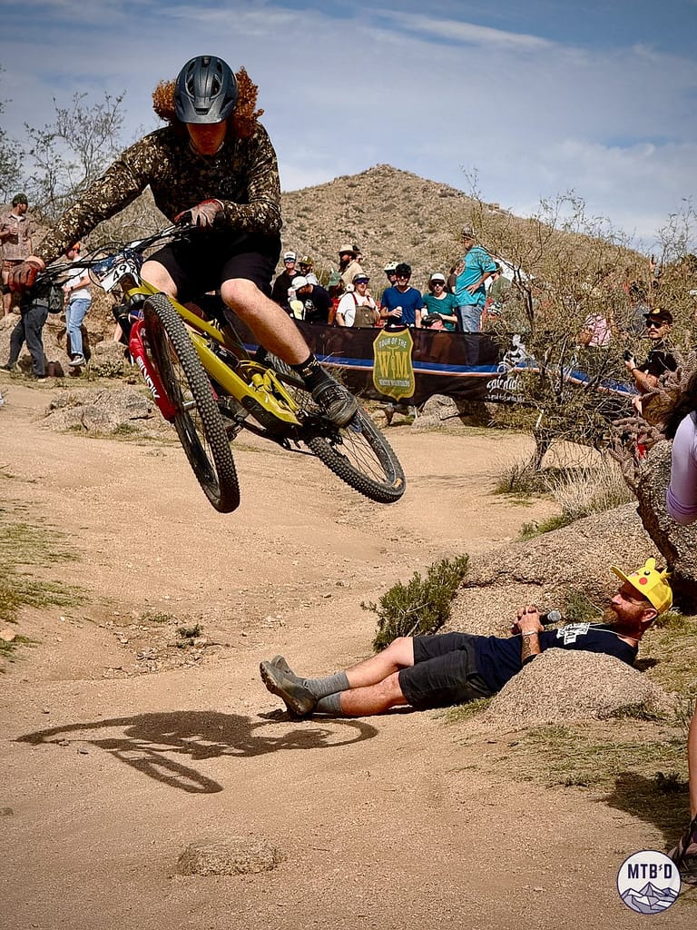 Mountain biker launching off the kicker jump over a man lying on the ground at 24 Hours in the Old Pueblo near Tucson, Arizona, with spectators watching from the trailside